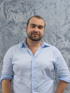 Adam is a bearded man with close-cropped hair standing in front of a light blue abstract artwork.