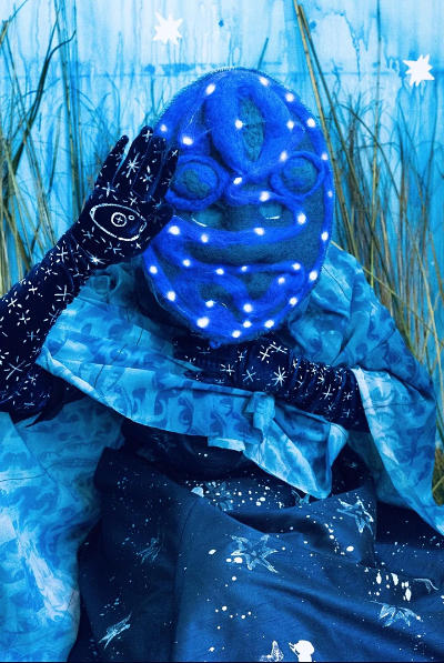 Masked figure all in blue celestial patterns waving with their gloved right hand. 